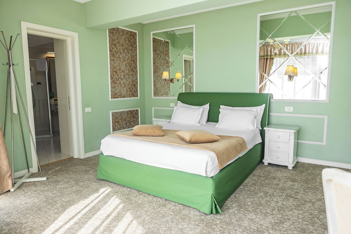 A bedroom with a green bed, white bedding, a nightstand, a mirror, and a window with curtains.