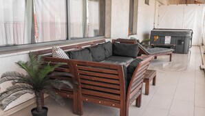 Un sofá de madera con cojines, un banco y una planta en maceta en una habitación con ventanas amplias y suelo de baldosas.
