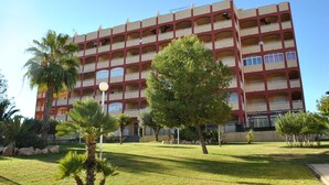 Um hotel com vários andares, fachada em vermelho e bege, varandas em cada andar e um gramado bem cuidado com palmeiras.