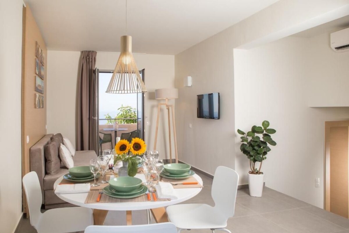 A dining area with a round table set for four, green plates, and a vase with sunflowers. There is a sofa and a TV in the background.