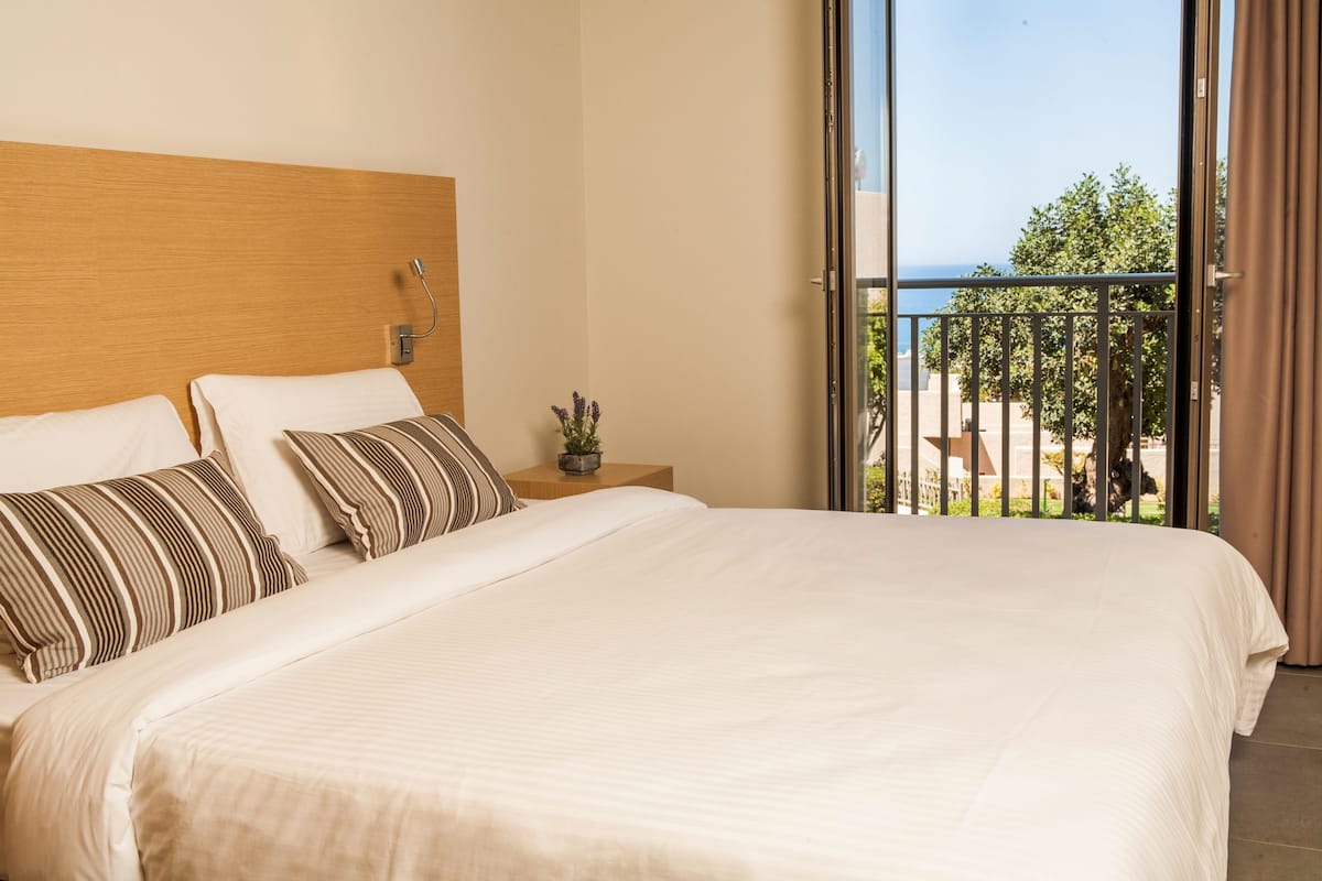 A hotel room with a large bed, a wooden headboard, a small bedside table with a plant, and a balcony with a view of the sea.