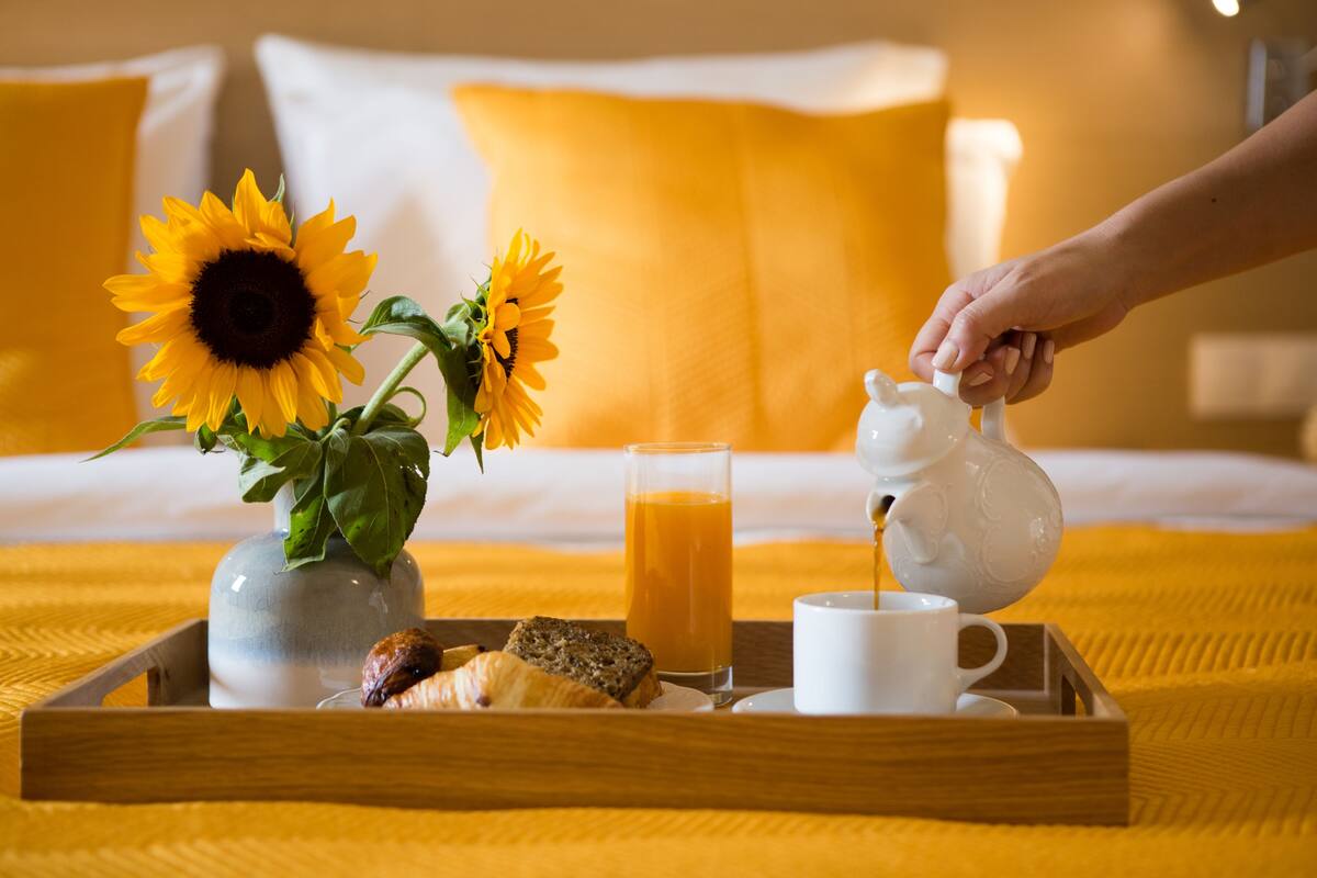 A breakfast tray with a vase of sunflowers, a glass of orange juice, a teapot, a cup, and a croissant on a wooden tray.