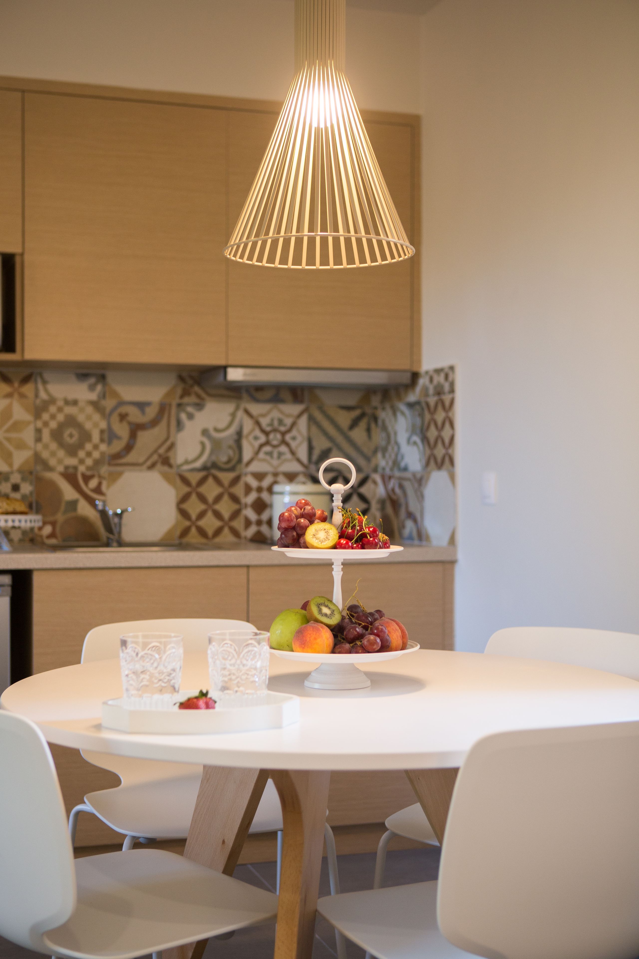 A modern kitchen with a white dining table, two chairs, a fruit display, and a hanging light fixture.