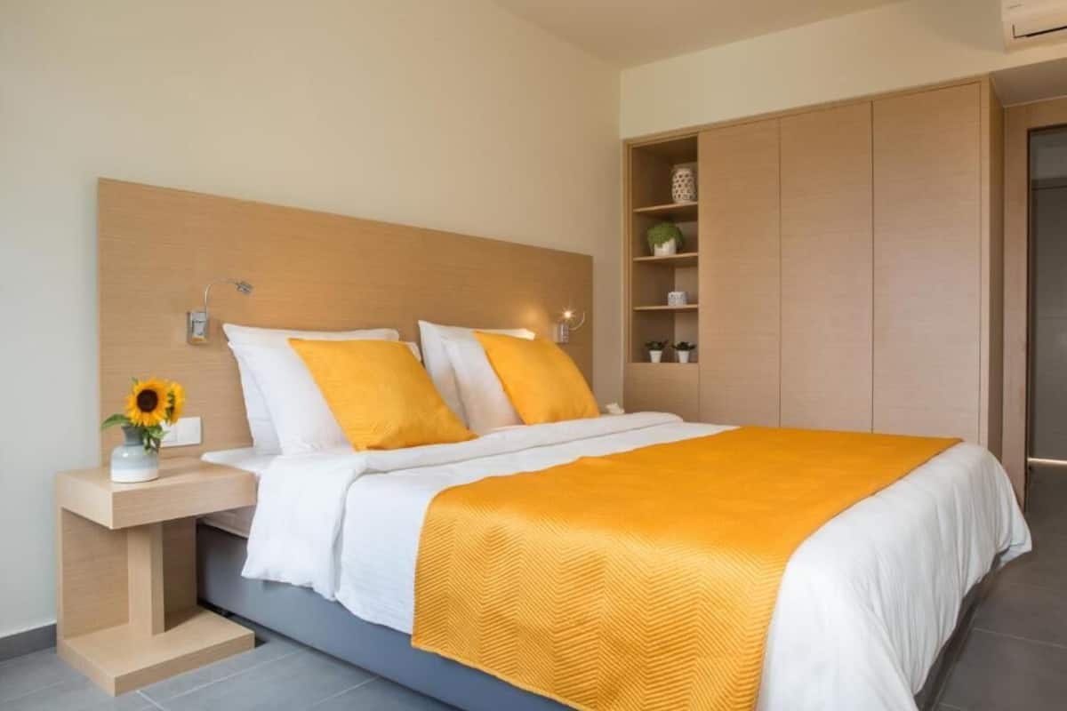 A modern bedroom with a wooden headboard, a bed with white and yellow bedding, a bedside table with a vase of sunflowers, and built-in shelving with decorative items.