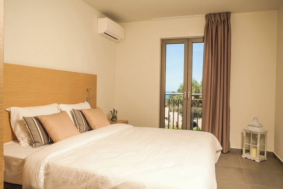 A hotel room with a large bed, a wooden headboard, a nightstand with a plant, a window with curtains, and a balcony with a view.