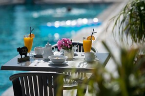 A dining table with breakfast items and drinks by a poolside, including a piggy bank and a flower vase. - Sundance Apartments and Suites (Hersonissos)