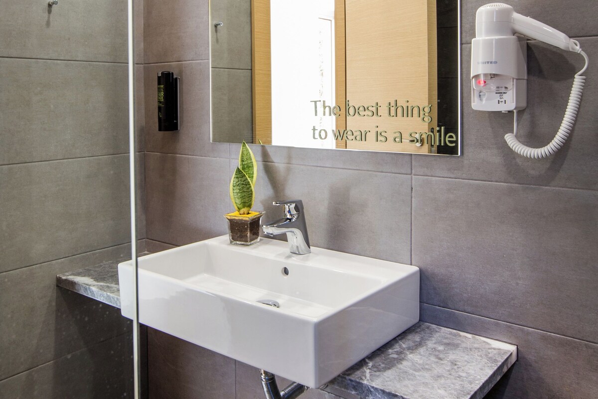 A modern bathroom with a large mirror, a white sink, a hairdryer, and a potted plant.