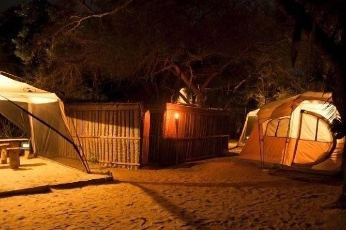 Um camping com duas tendas e uma estrutura de madeira, possivelmente um depósito ou espaço de armazenamento, à noite.