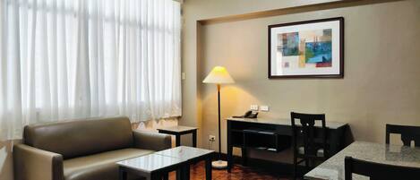 A hotel room with a sofa, a small table, a desk with a lamp, and a framed picture on the wall.