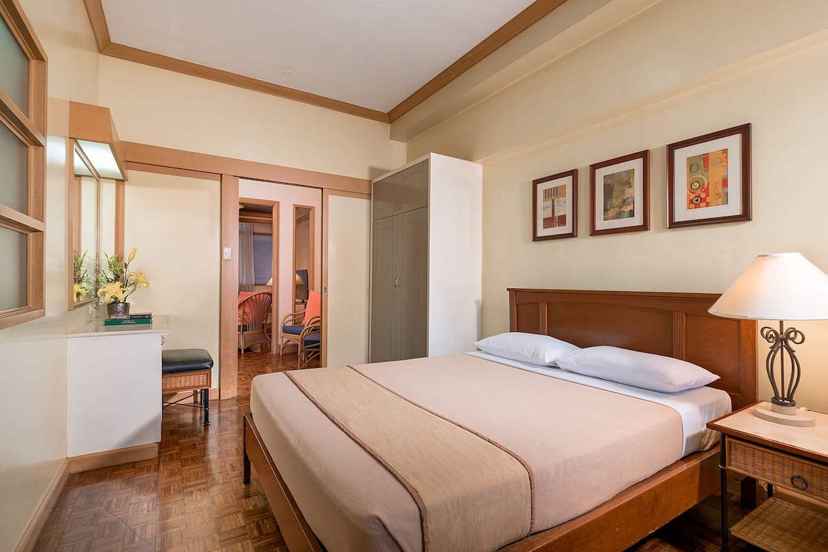 A hotel room with a bed, bedside table, lamp, framed pictures on the wall, a desk with a chair, and a mirror.