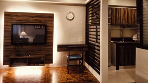 A modern living room with a flat-screen TV, a wooden entertainment unit, a dining area with a chair, and a kitchen with wooden cabinets. - Tropicana Suites (Manila)