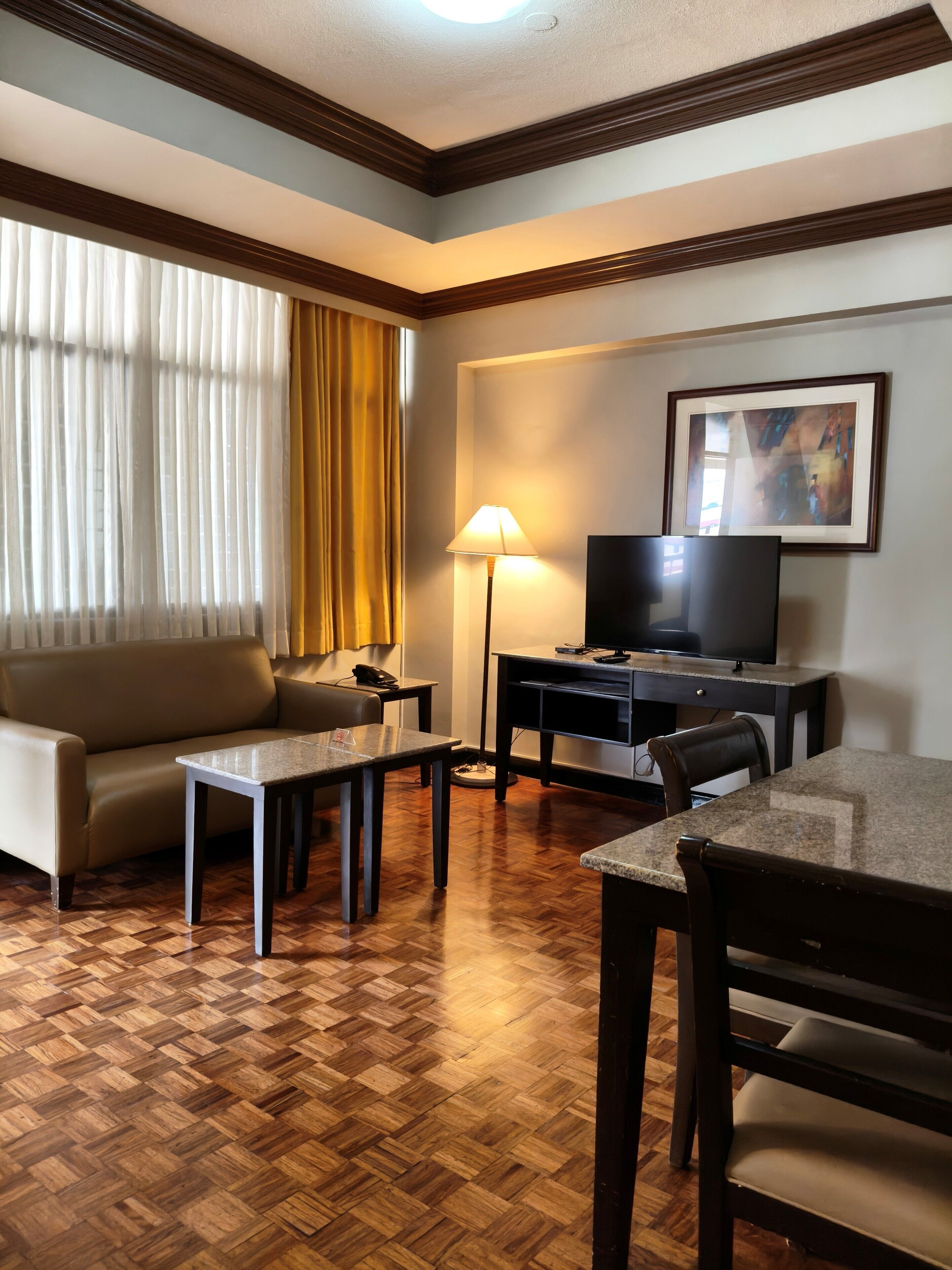 Habitación de hotel con suelo de madera en patrón de espiga, un sofá, una mesa de centro, un televisor sobre un mueble bajo y un cuadro en la pared.
