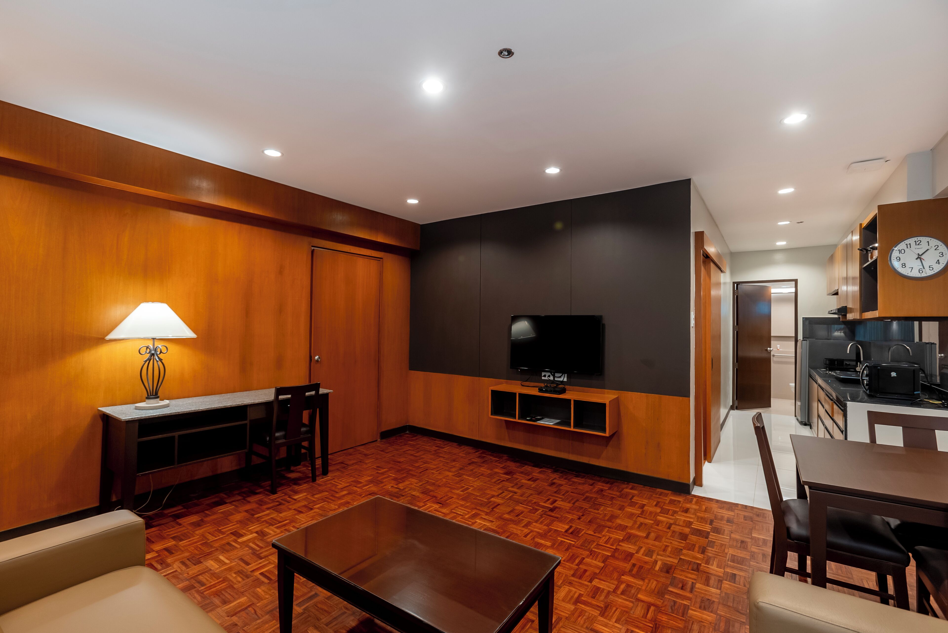 A modern living room with a wooden floor, a TV on a black wall, a dining area with a table and chairs, and a kitchen in the background.