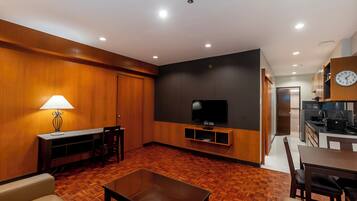 A modern living room with a wooden floor, a TV on a black wall, a dining area with a table and chairs, and a kitchen in the background.