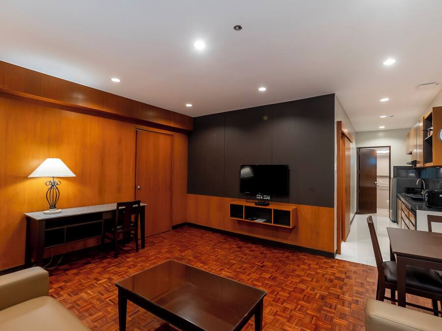 A modern living room with a wooden floor, a TV on a black wall, a dining area with a table and chairs, and a kitchen in the background.