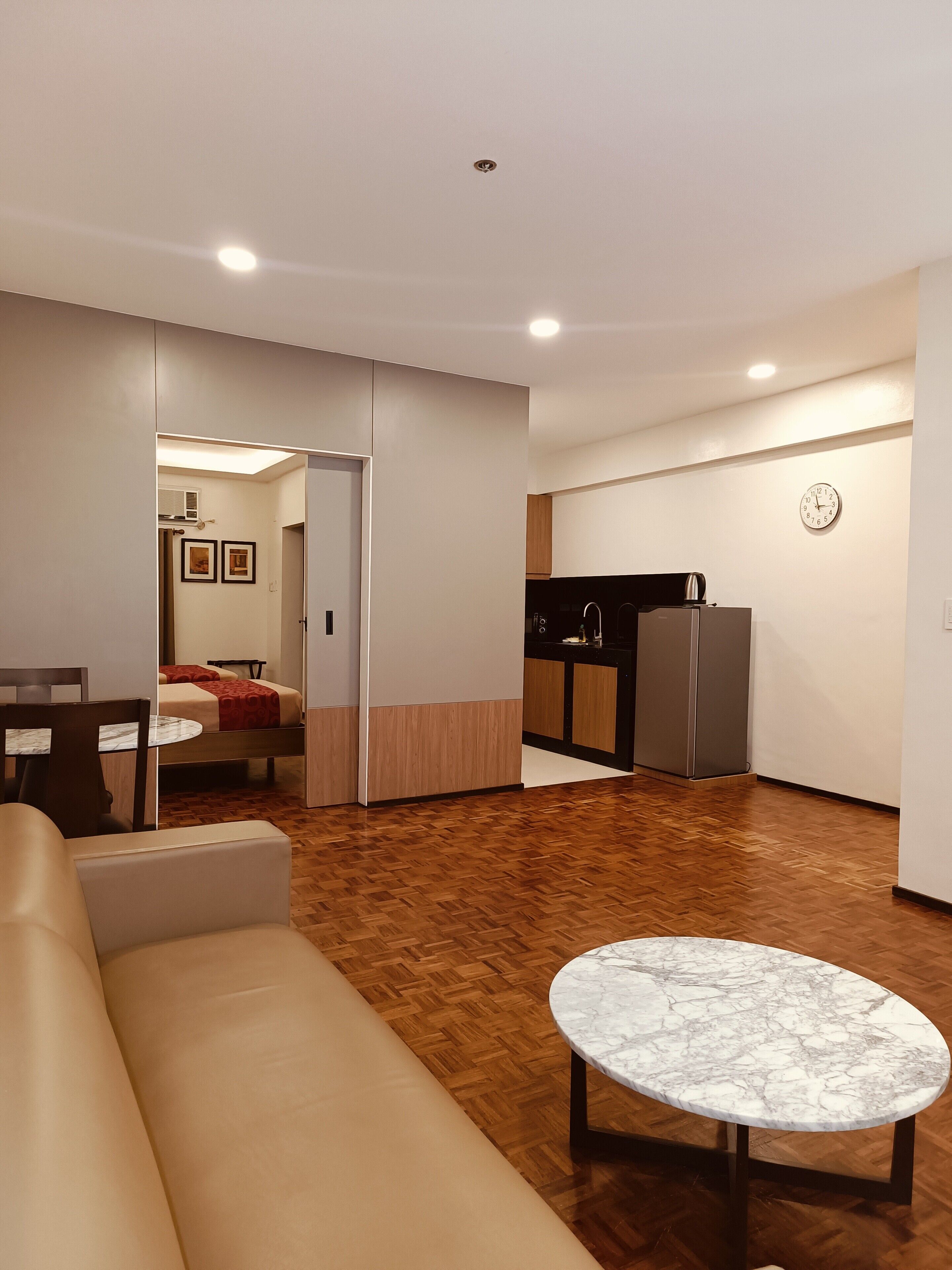 Una sala moderna con sofá, una mesa baja de mármol y suelo de madera. Cuenta con una kitchenette con refrigerador y un reloj en la pared.