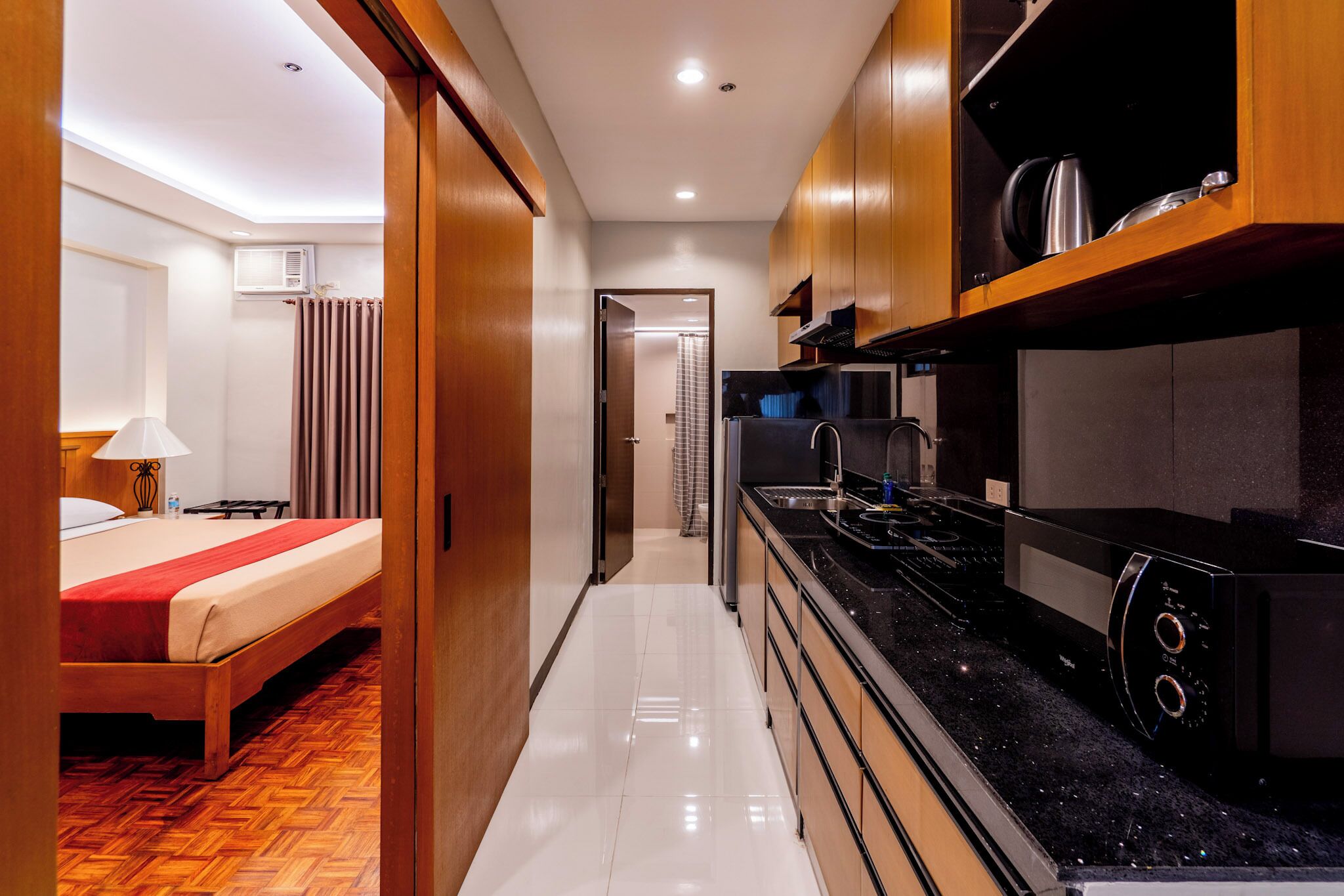 A hotel room with a bed, a small desk, a kitchenette with a stove and microwave, and a bathroom with a shower curtain.