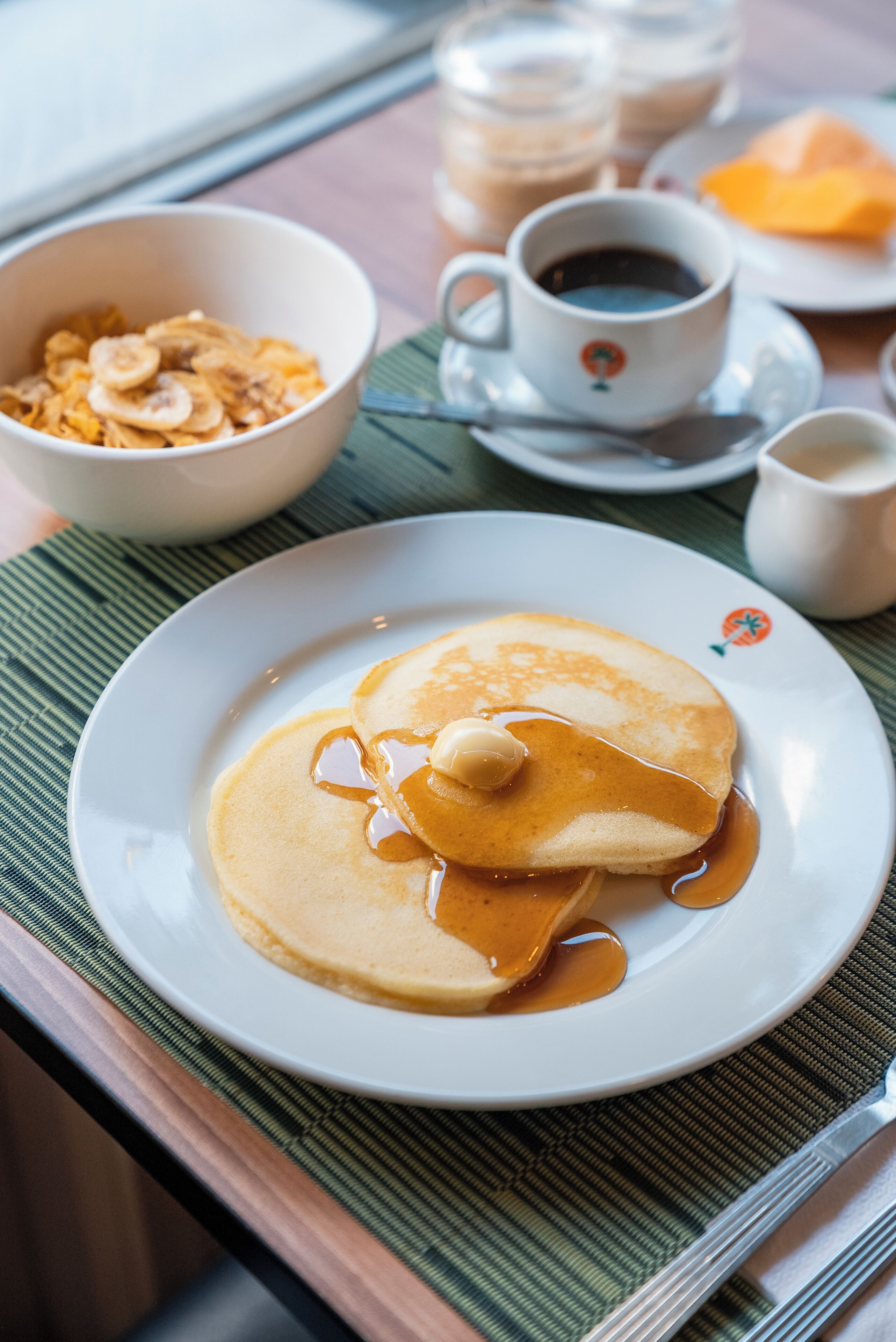 A breakfast setting with pancakes on a plate, a cup of coffee, and a bowl of cereal.