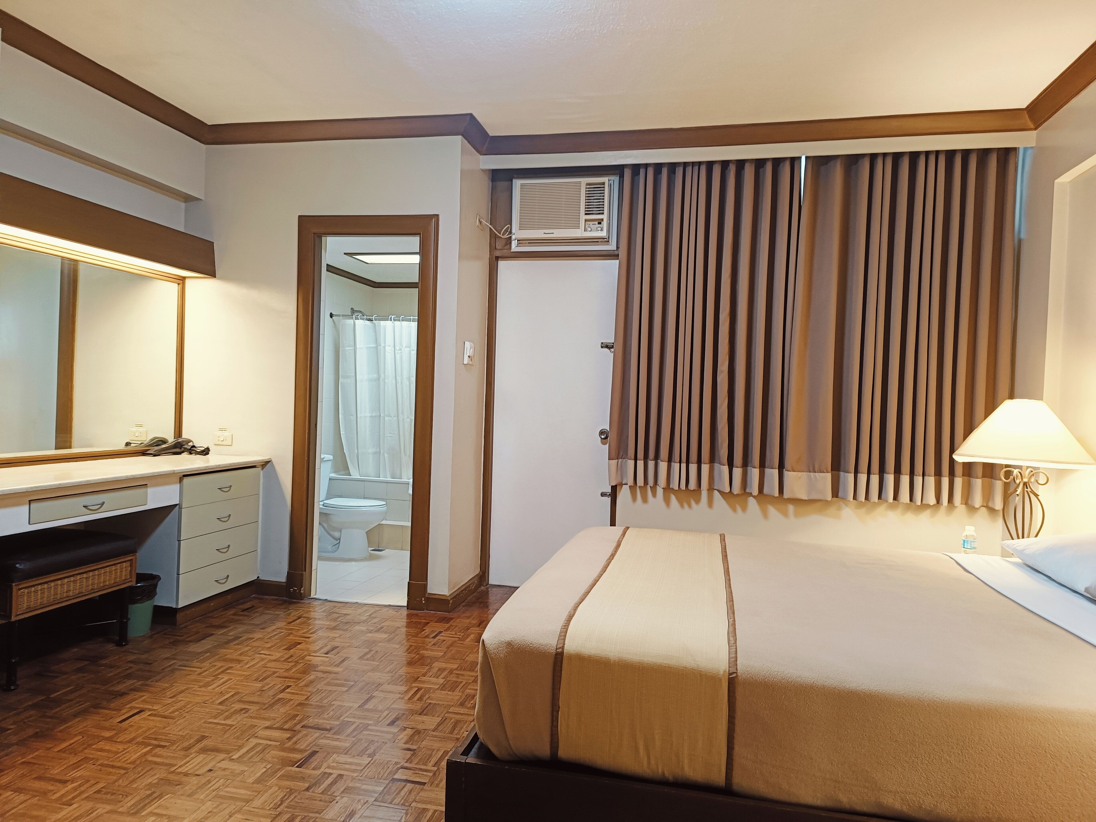 Une chambre d’hôtel comprenant un lit, un bureau avec des tiroirs, un miroir, un appareil de climatisation et une fenêtre avec des rideaux.