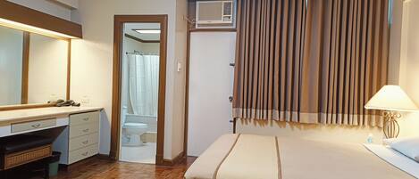 Habitación de hotel con cama, escritorio con cajones, espejo, unidad de aire acondicionado y ventana con cortinas.