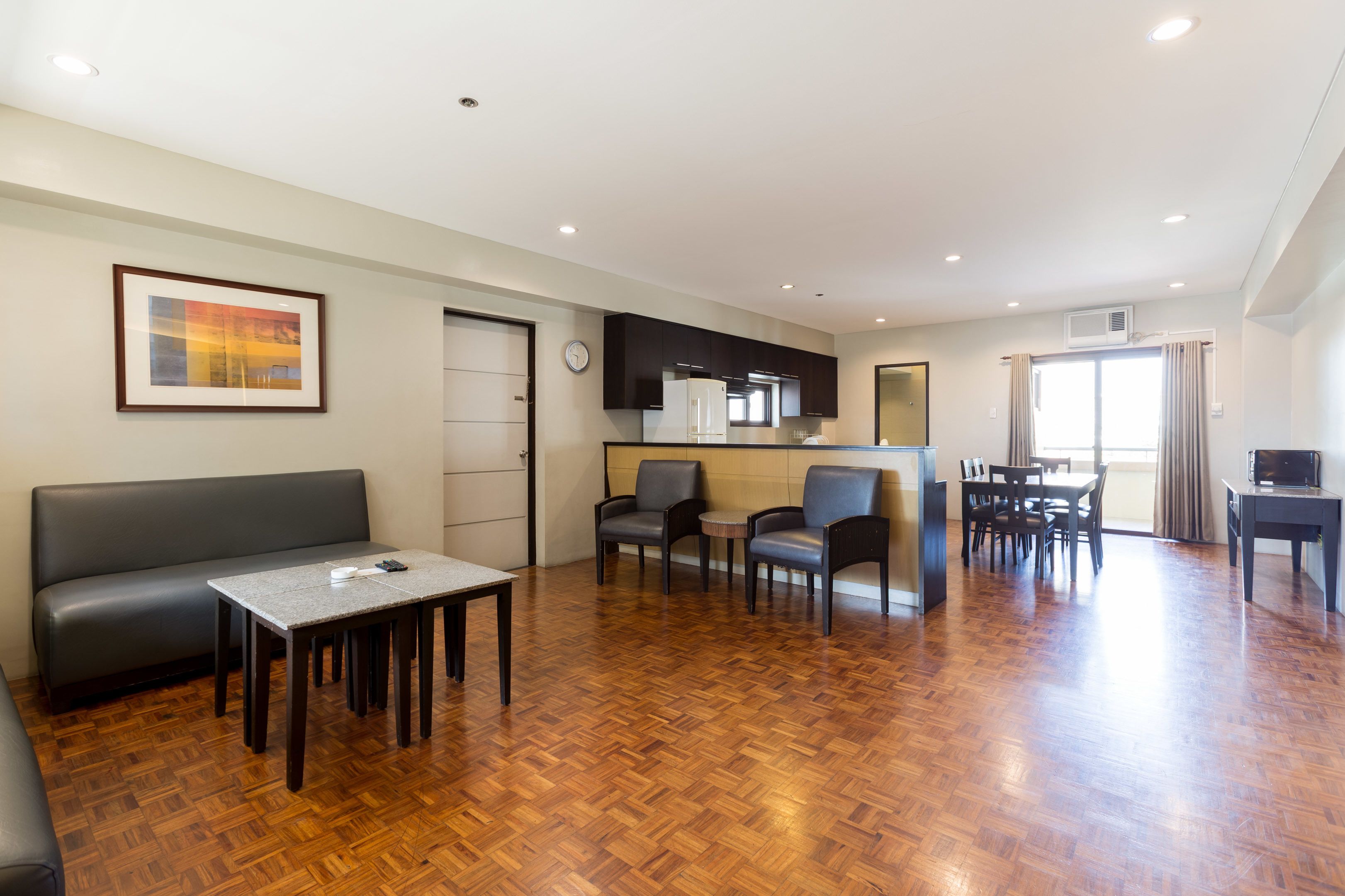A modern living room with a sofa, a dining table, and chairs. There is a kitchen area with cabinets and a refrigerator.