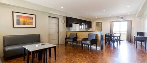 A modern living room with a sofa, a dining table, and chairs. There is a kitchen area with cabinets and a refrigerator.