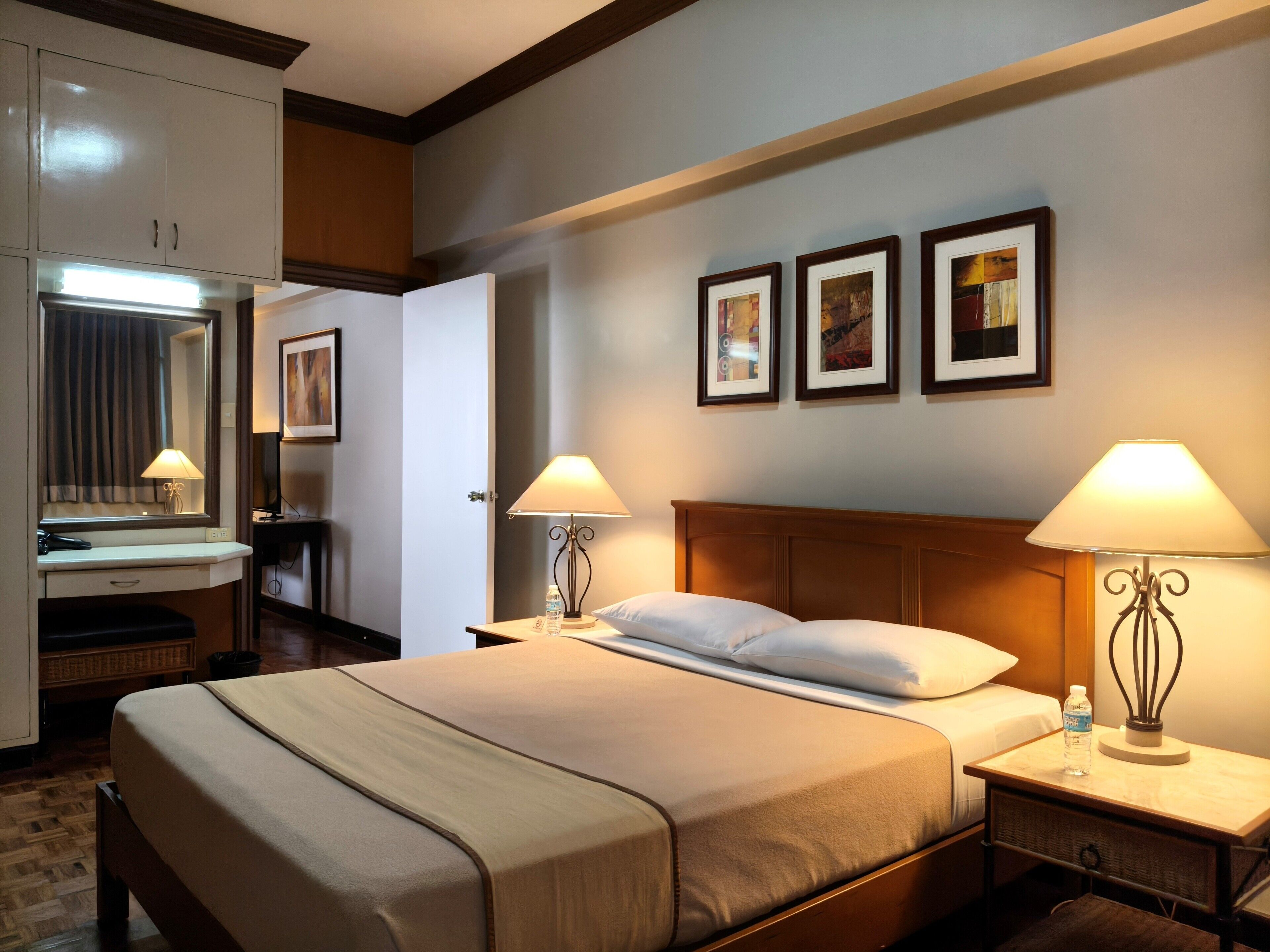 A hotel room with a large bed, bedside tables with lamps, a dresser, a mirror, and framed pictures on the wall.