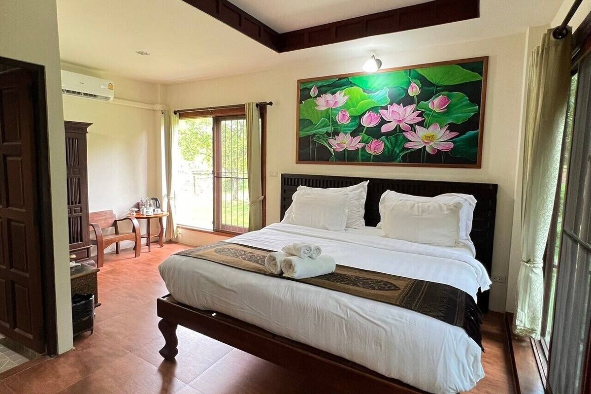 Habitación de hotel con una cama grande, un cuadro de flores de loto, un banco de madera y vistas al exterior a través de una puerta.