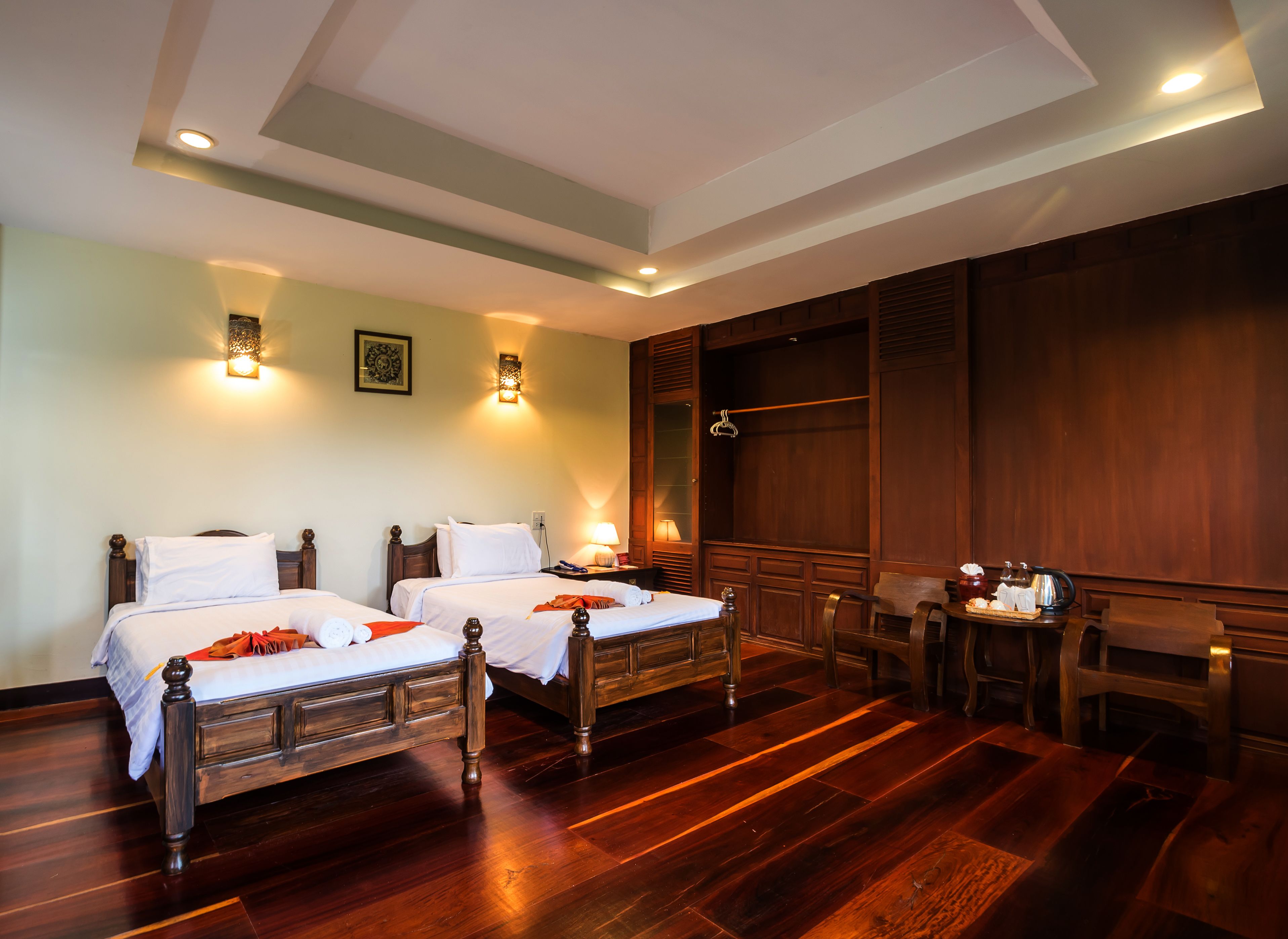 Una habitación de hotel con dos camas de madera, un armario de madera, una mesita con una lámpara y una silla de madera.