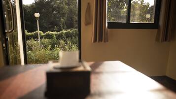 A room with a table, a window with curtains, and a view of greenery outside.