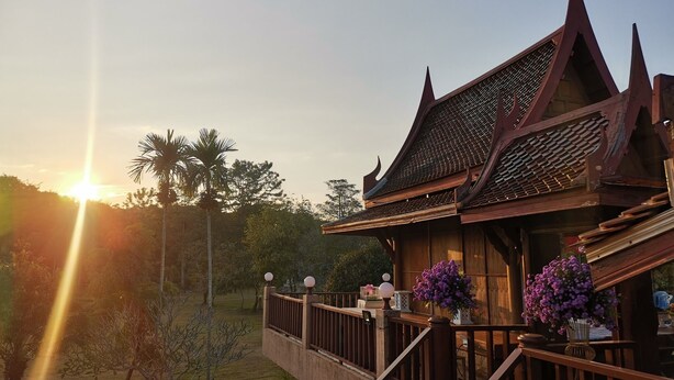 Un bâtiment traditionnel en bois, doté d’un balcon, entouré de verdure et de palmiers, au coucher du soleil.