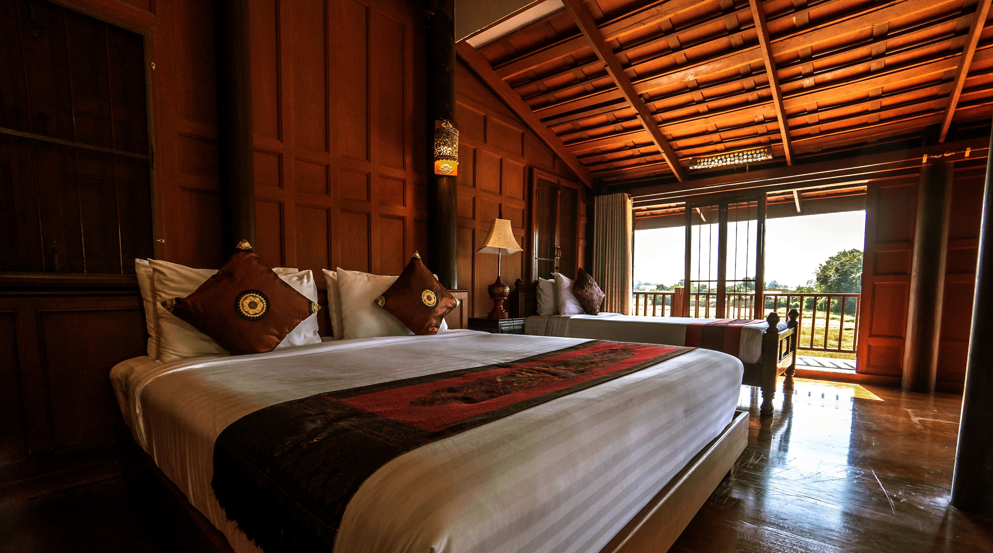 A large bed with multiple pillows, a nightstand with a lamp, and a view of the outdoors through a large window.