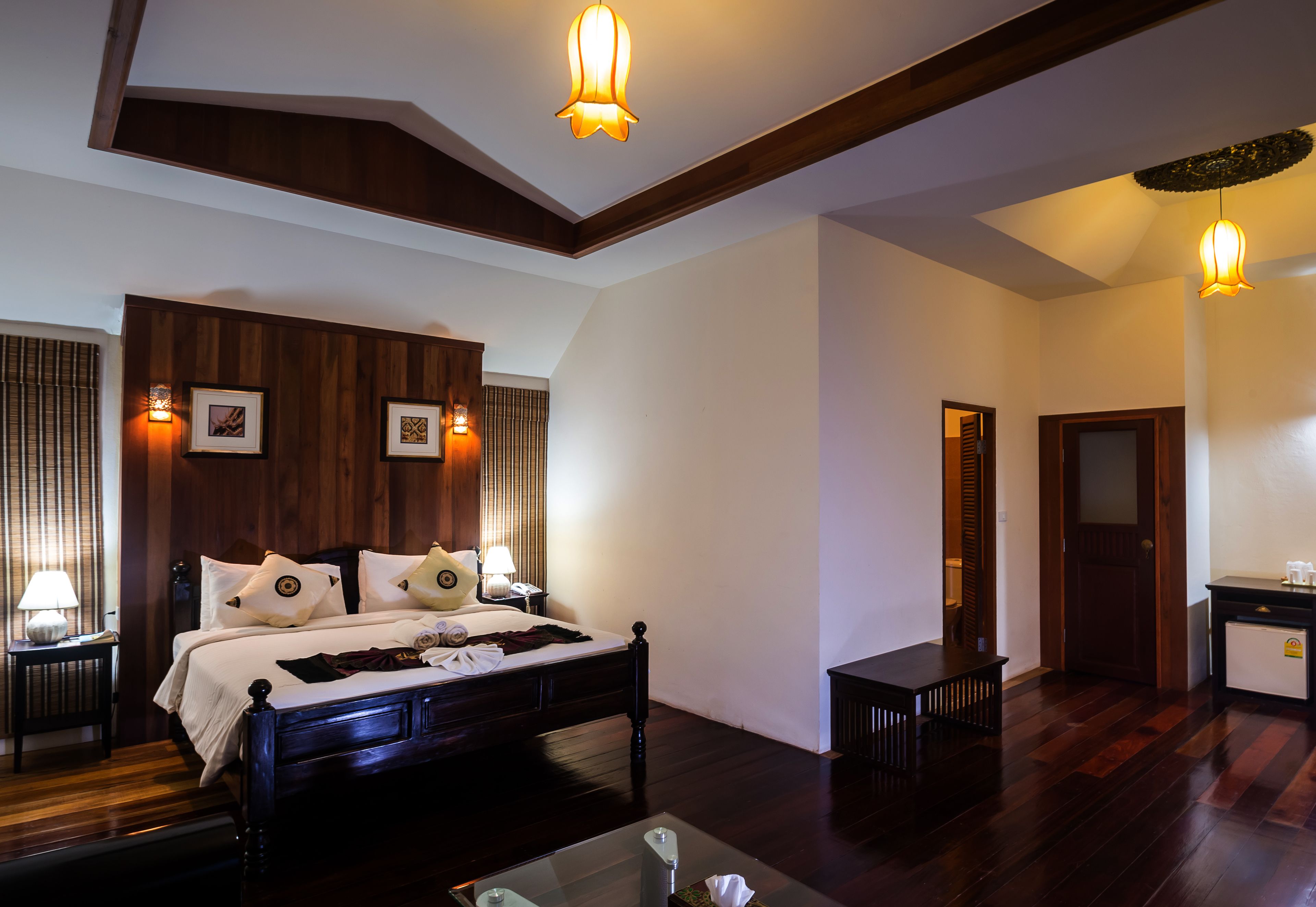 A bedroom with a wooden bed, bedside tables with lamps, a glass coffee table, and a door leading to another room.