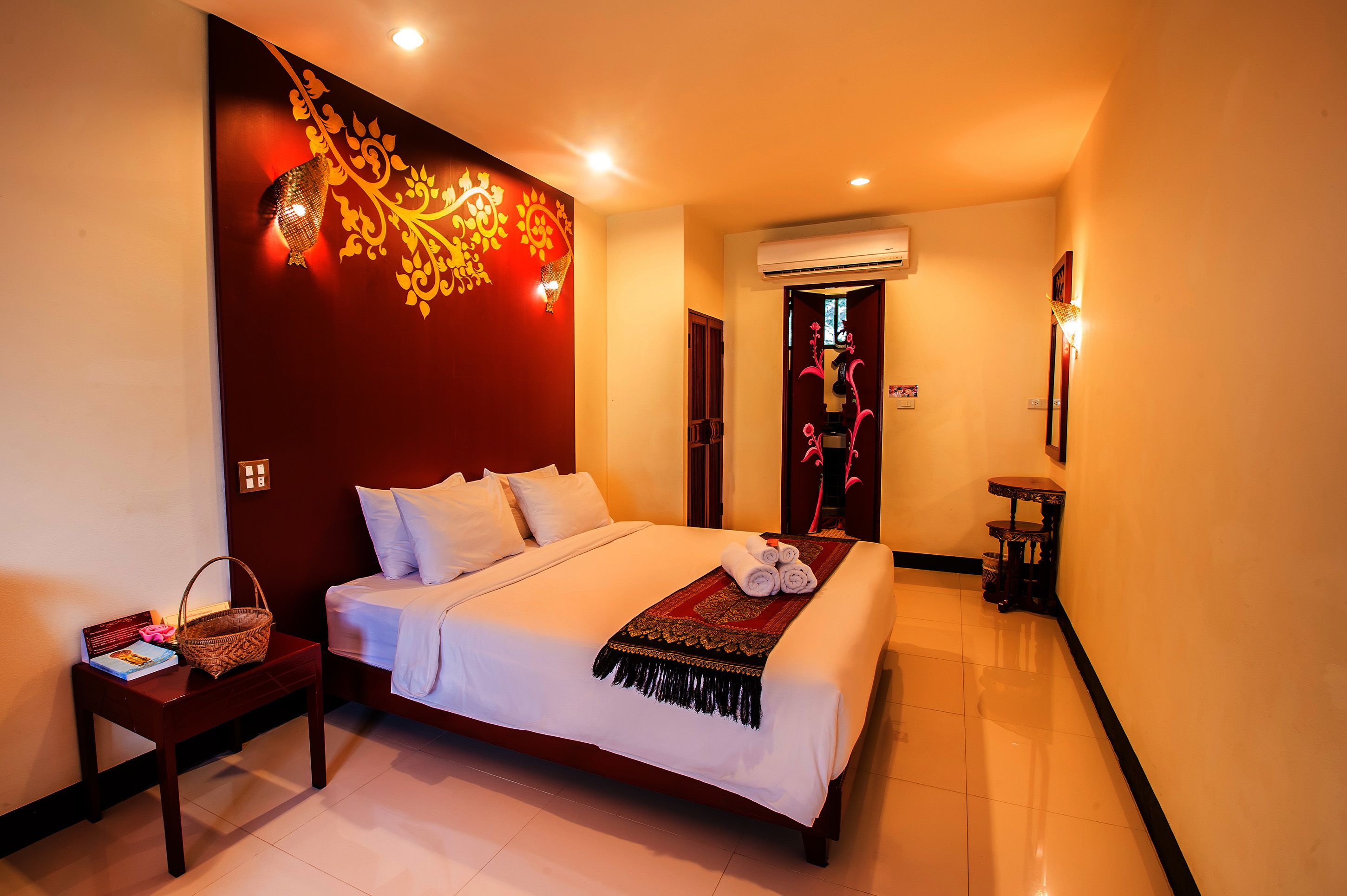 A neatly made bed with white linens, a decorative wall with a golden design, and a small table with a basket and book.