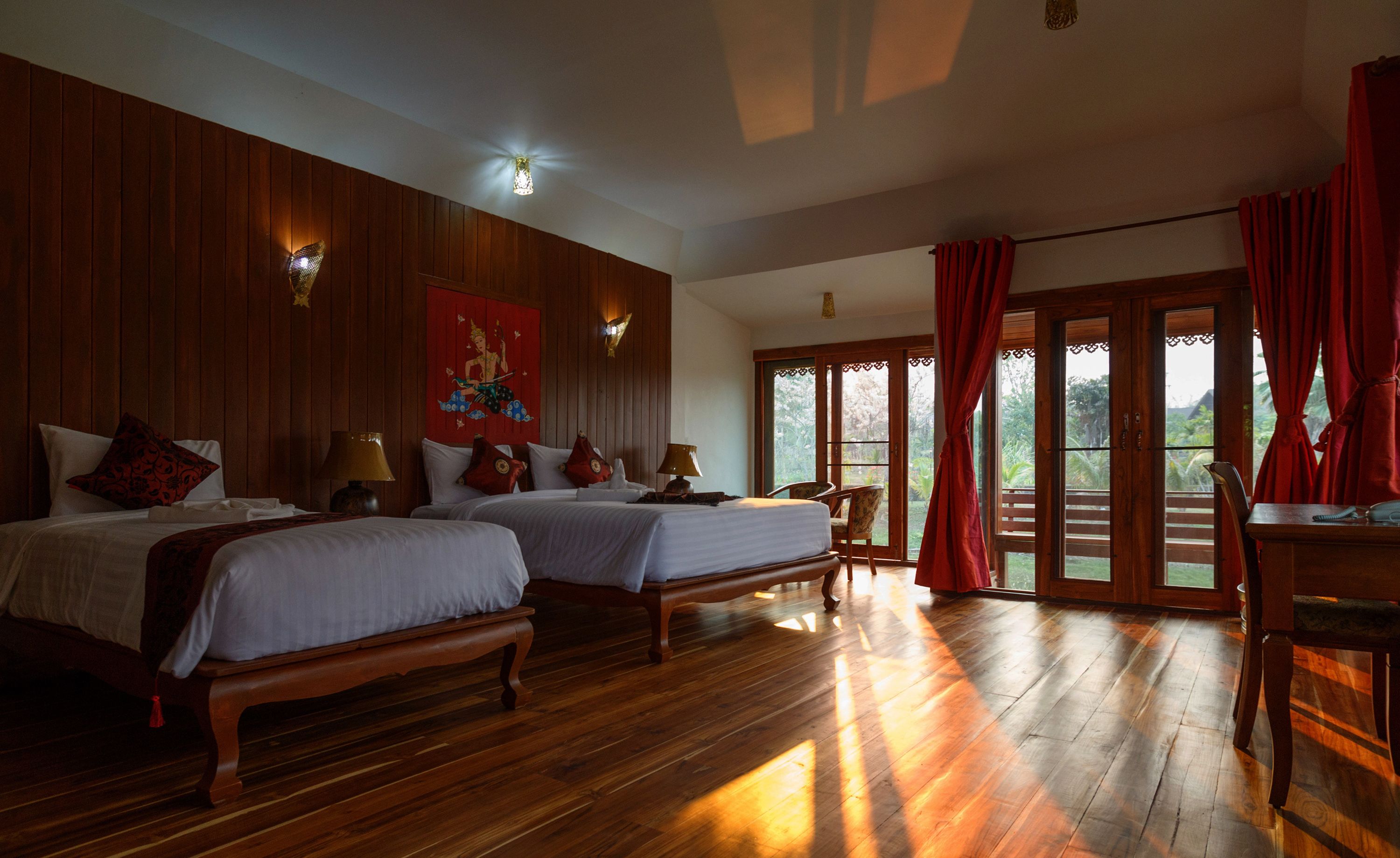 A hotel room with two beds, wooden flooring, a red curtain, and a view of greenery outside.