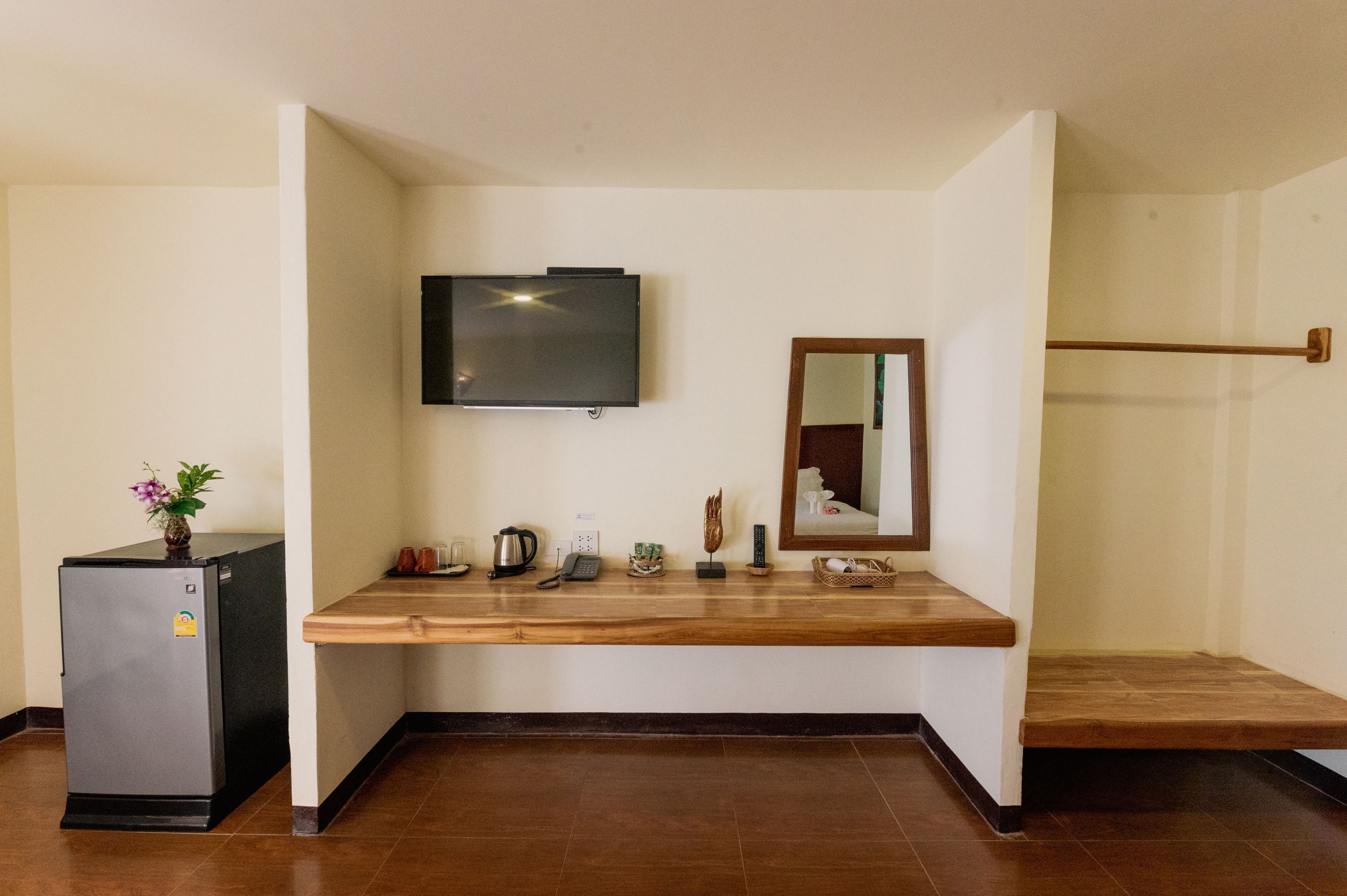 A hotel room with a wooden counter, a television mounted on the wall, a refrigerator, a small table with a plant, a coffee maker, a phone, a basket, and a mirror.