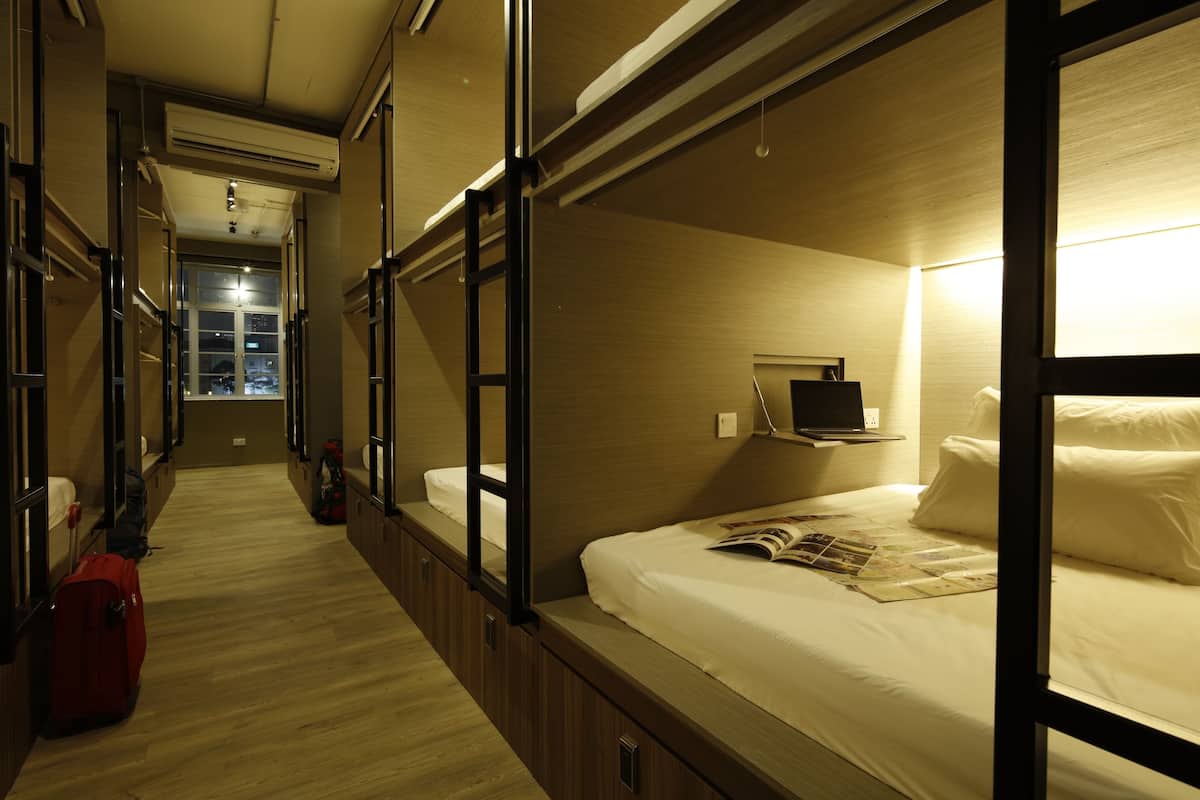 Ein Hotelzimmer mit Etagenbett, Nachttisch mit Lampe, Fenster mit Blick auf die Stadt und einem Koffer auf dem Boden.
