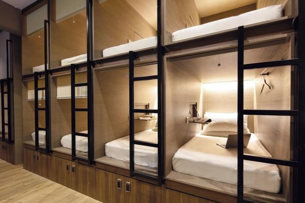 A row of bunk beds with wooden frames and white bedding, each with a small shelf and a hanging item.