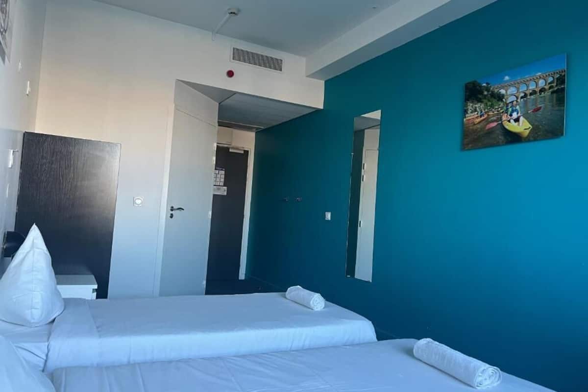 Habitación de hotel con dos camas, una pared de acento color verde azulado, un cuadro de un puente y una puerta que da a otra habitación.