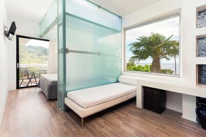 A modern room with a glass partition, a bed, a small table, a TV, and a view of the outdoors. - Kamari Beach Hotel (Thasos)