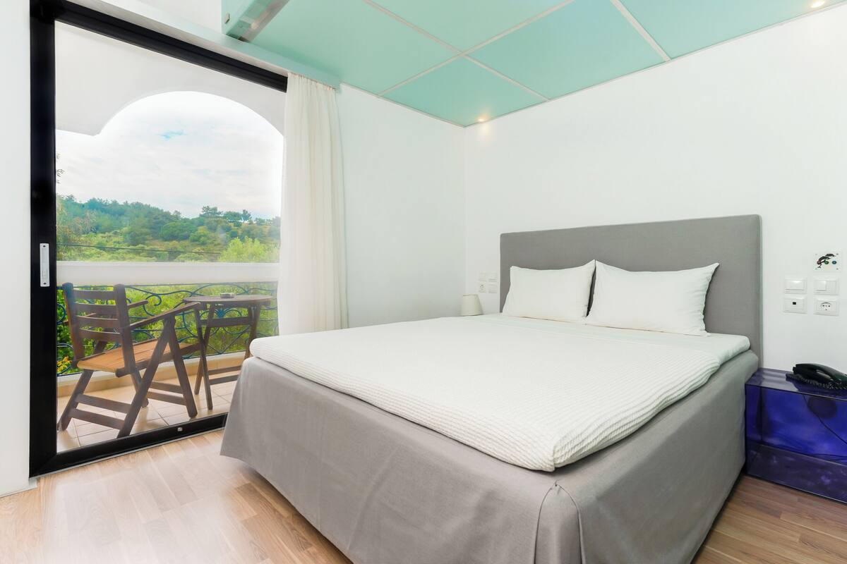 Ein Schlafzimmer mit Bett, Nachttisch, Lampe, Telefon und Blick auf einen Balkon mit Stühlen.