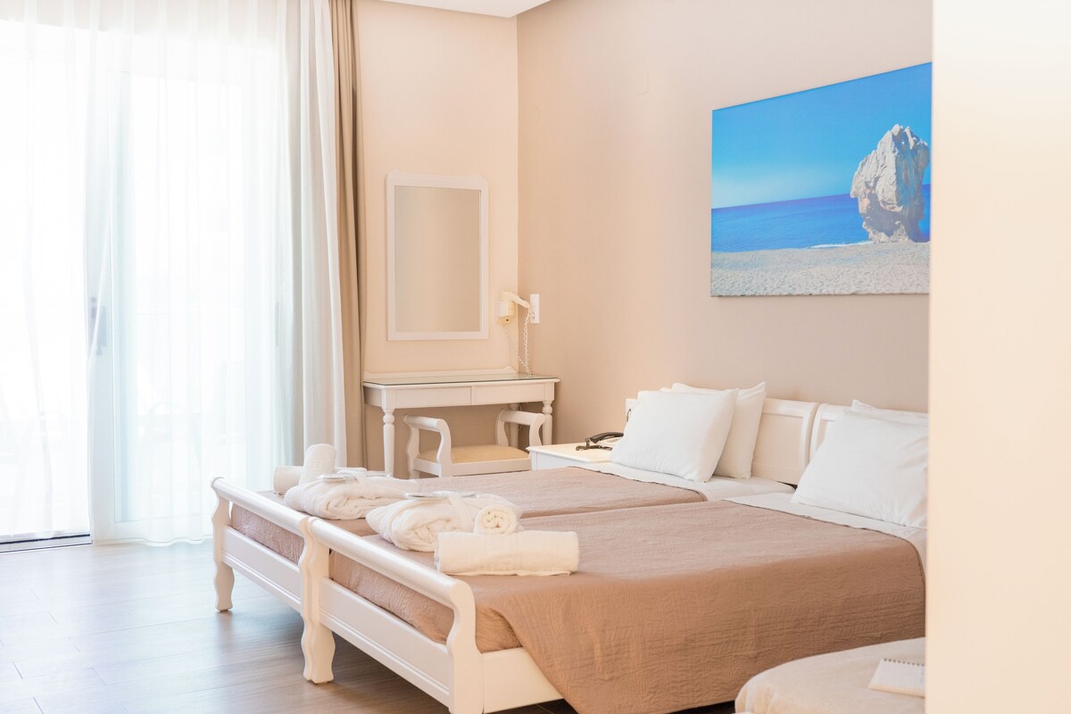 Una habitació d'hotel amb un llit, una taula de treball, una cadira i un quadre que representa una escena de platja.