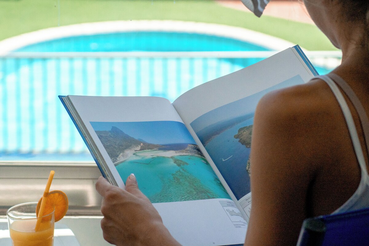 Una persona llegint un llibre al costat d'una piscina, amb una beguda a prop.