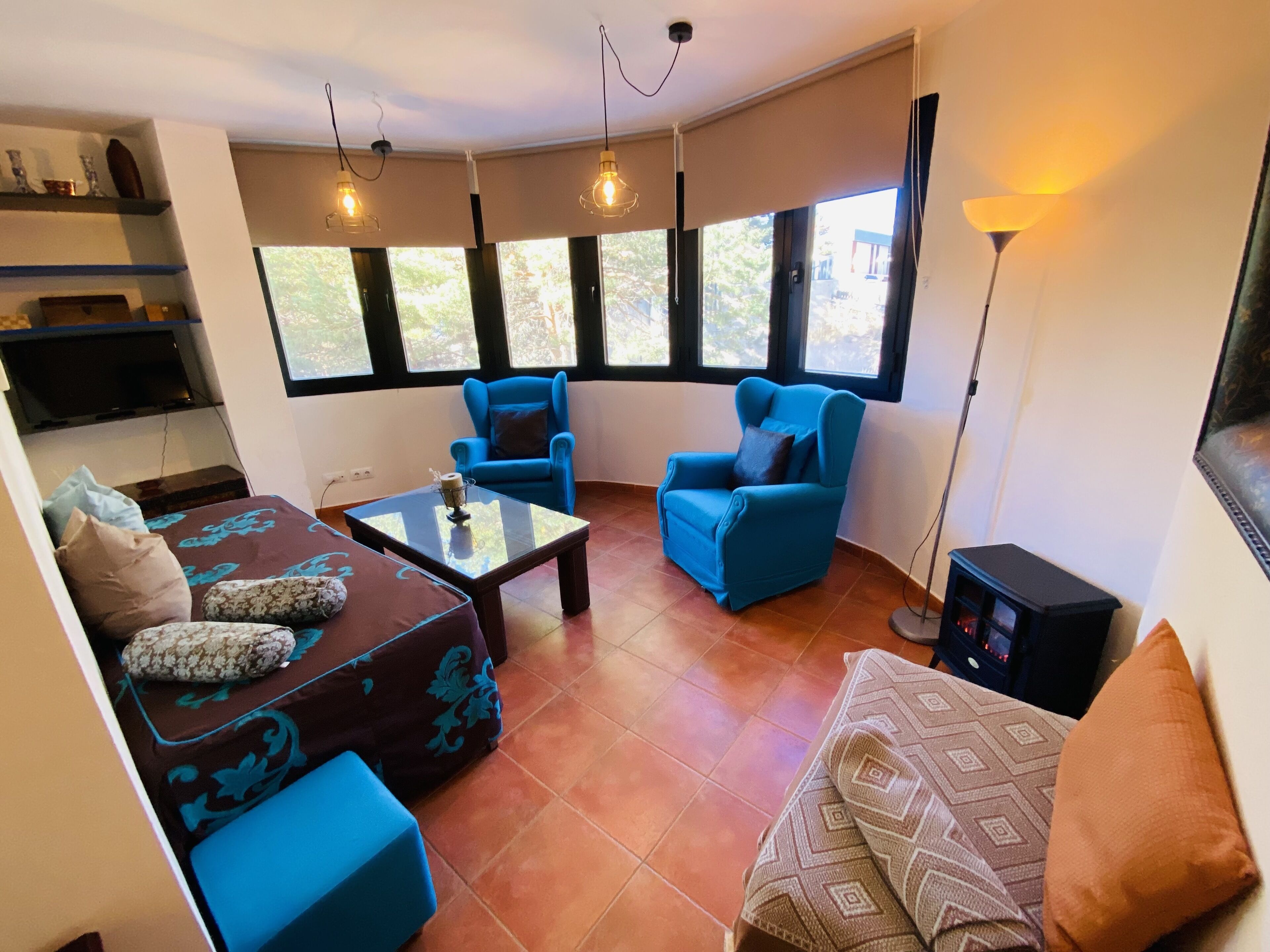 Una sala de estar con dos sillones azules, un sofá, una mesa de centro de vidrio y un televisor sobre una estantería.