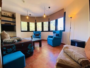A living room with a sofa, two armchairs, a coffee table, and a TV. There are windows with blinds, a ceiling light, and a floor lamp. - Snow Comfort Edificio Monte Gorbea (Monachil)