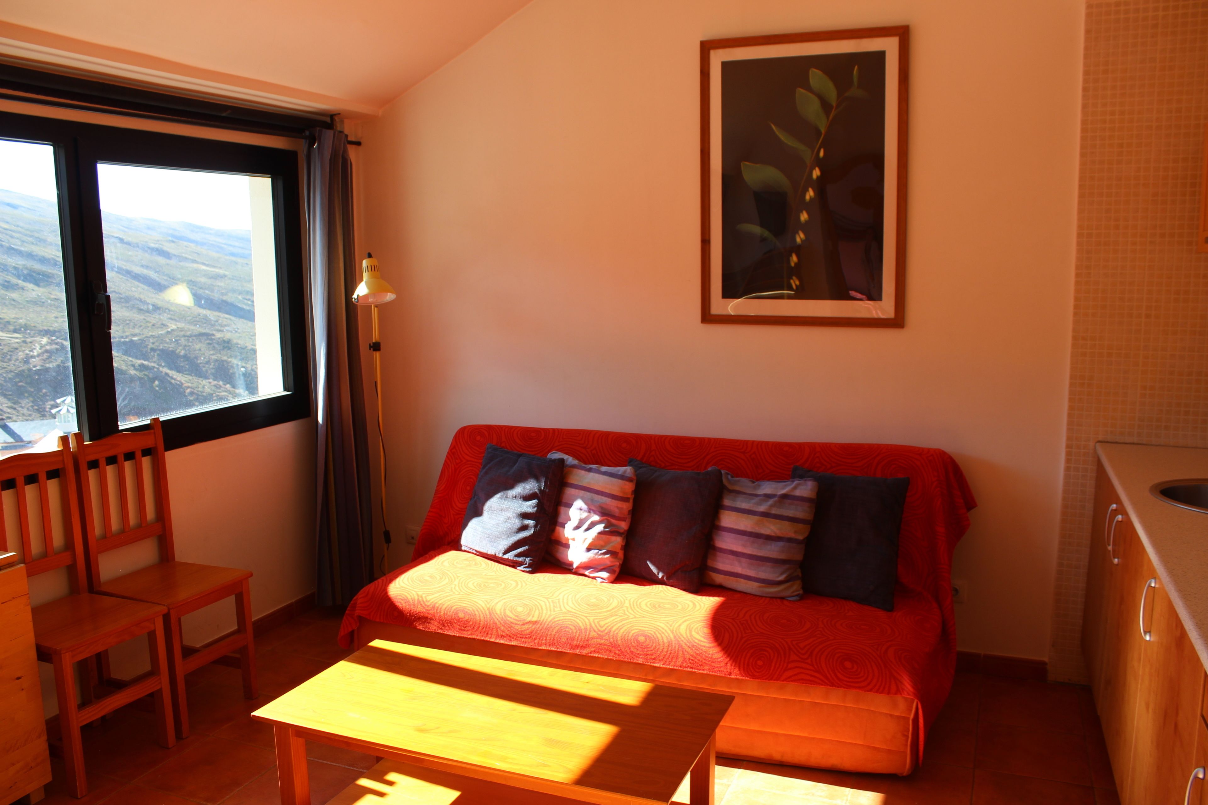 A cozy room with a red sofa, a wooden table, a chair, and a large window offering a view of the landscape.