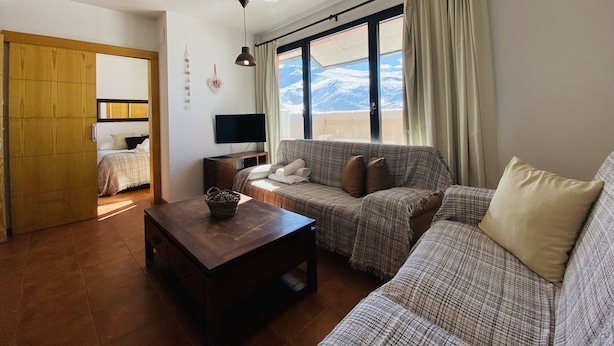 A cozy living room with a sofa, a coffee table, and a TV. There is a window with a view of mountains. - Snow Comfort Edificio Monte Gorbea (Monachil)