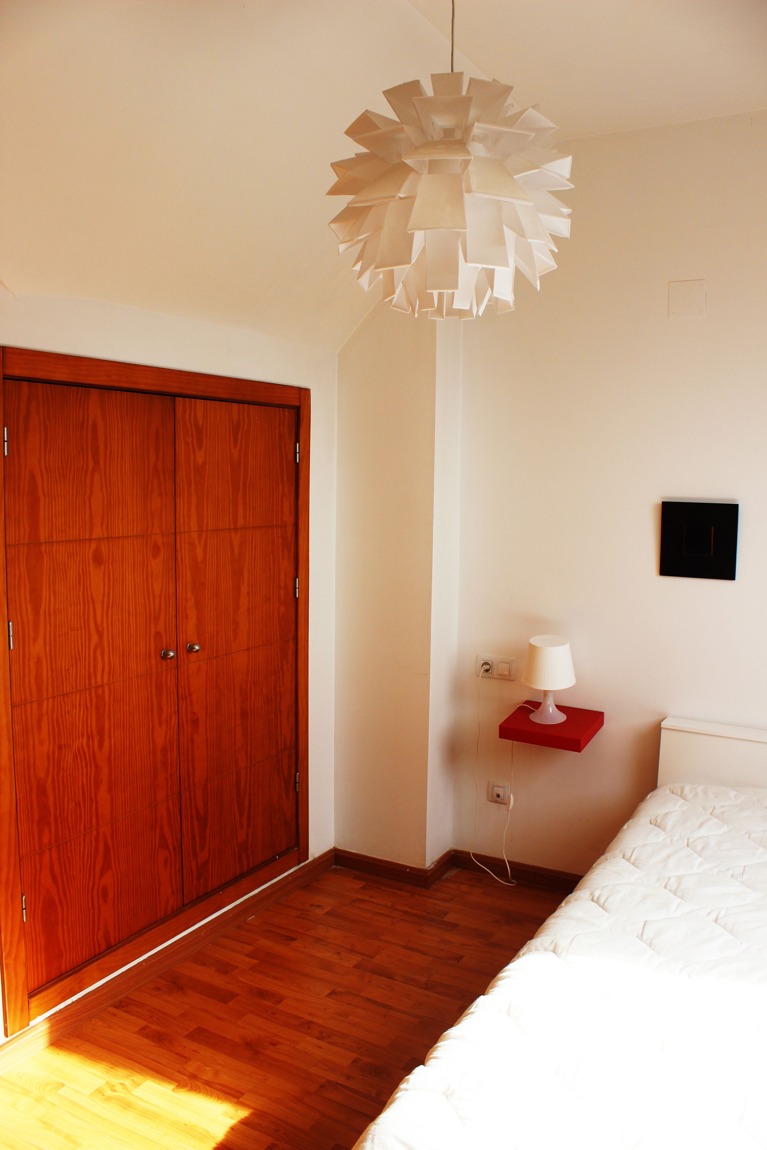 Un dormitorio con un armario de madera, una cama blanca, una mesita de noche roja con una lámpara y una lámpara en el techo.