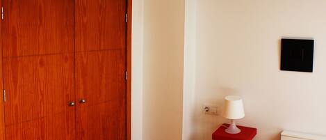 Un dormitorio con un armario de madera, una cama blanca, una mesita de noche roja con una lámpara y una lámpara en el techo.