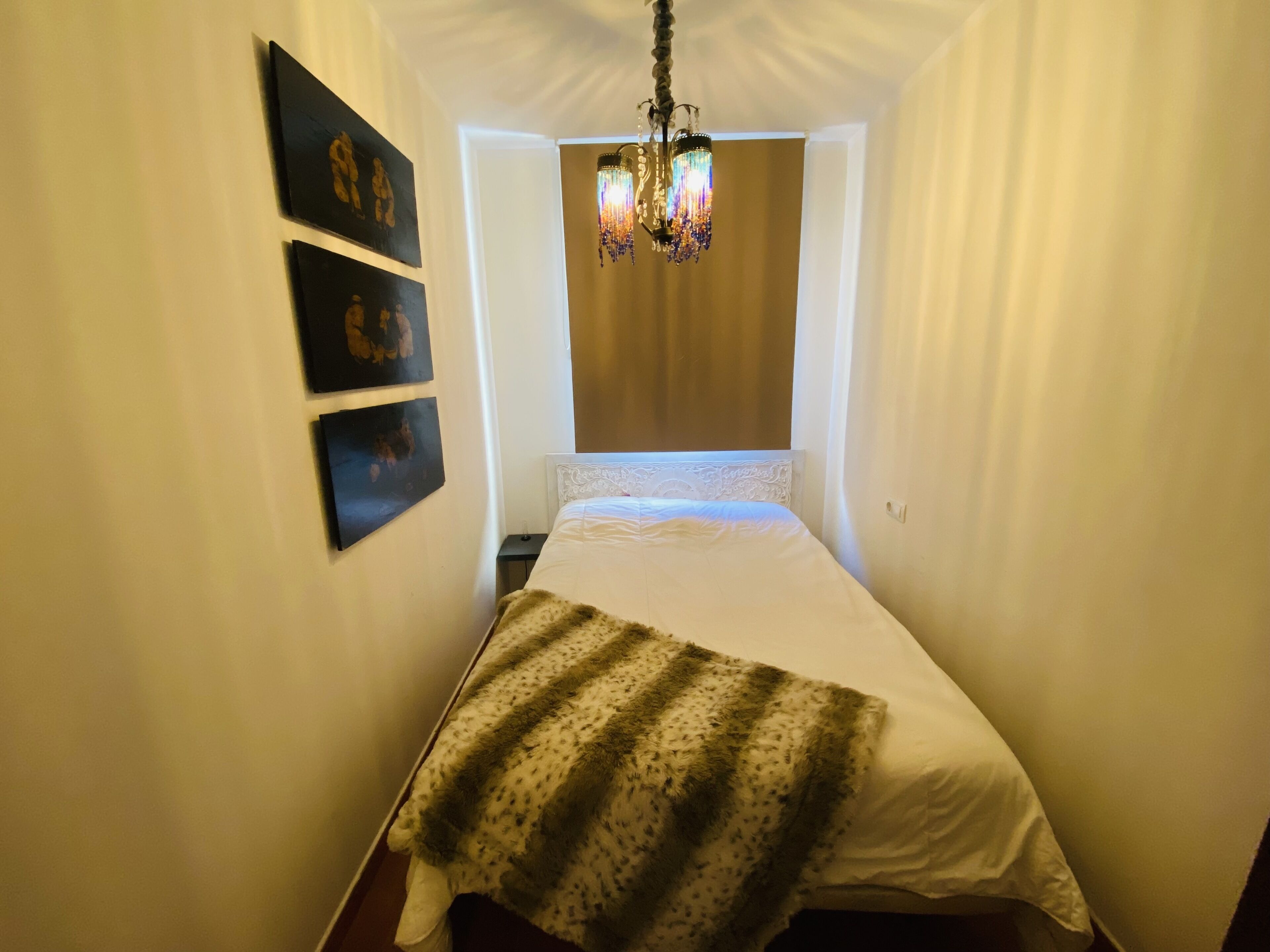 Habitació estreta amb un llit, un penjador de cristall i tres quadres amb marc a la paret.